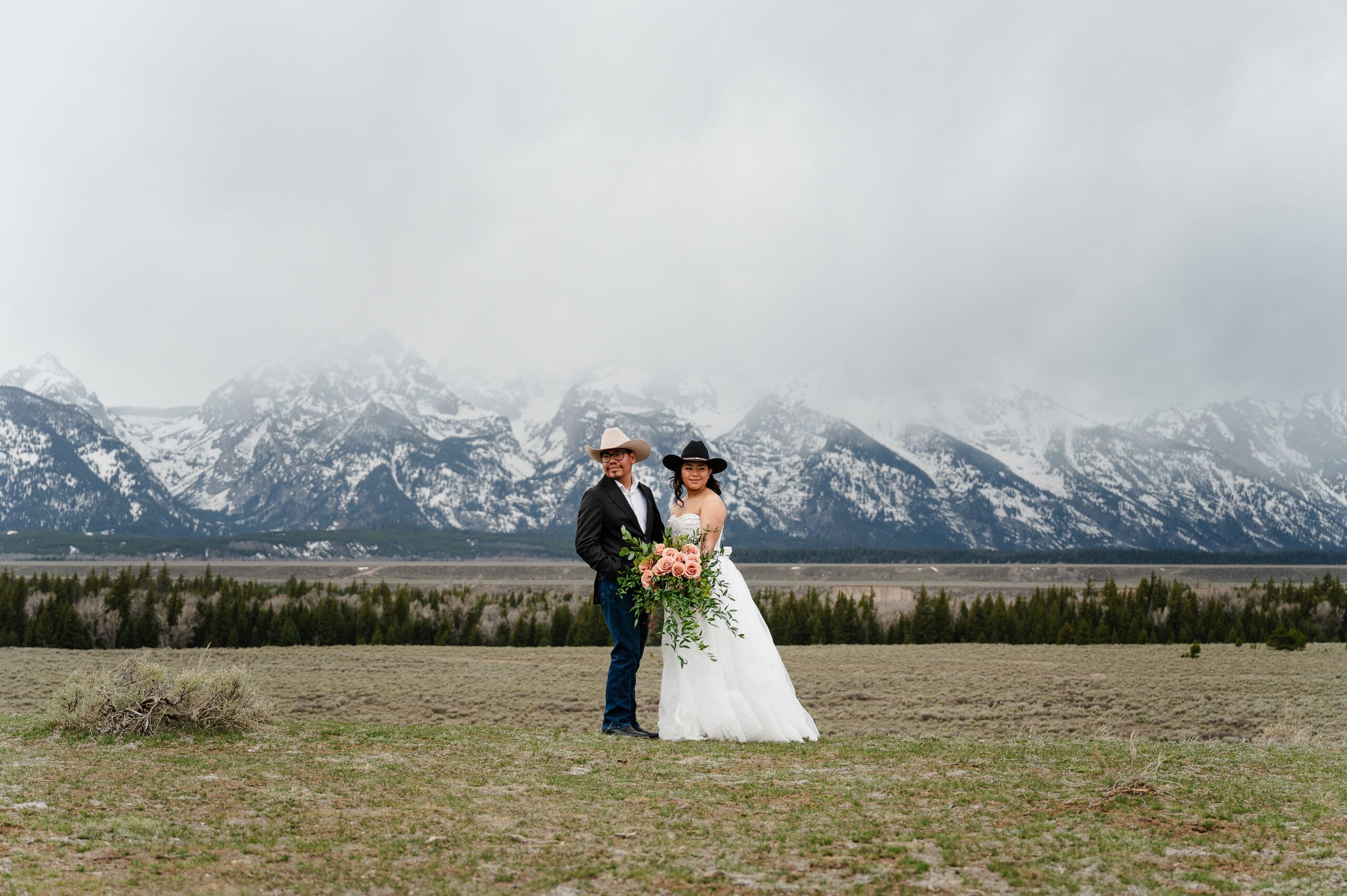 Elopement couple in wedding suit and dress with cowboy hats on at Glacier View Turnout in Grand Teton National Park.